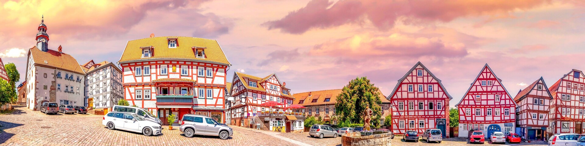 A cobblestone street lined with traditional half-timbered houses, a statue, and parked cars.