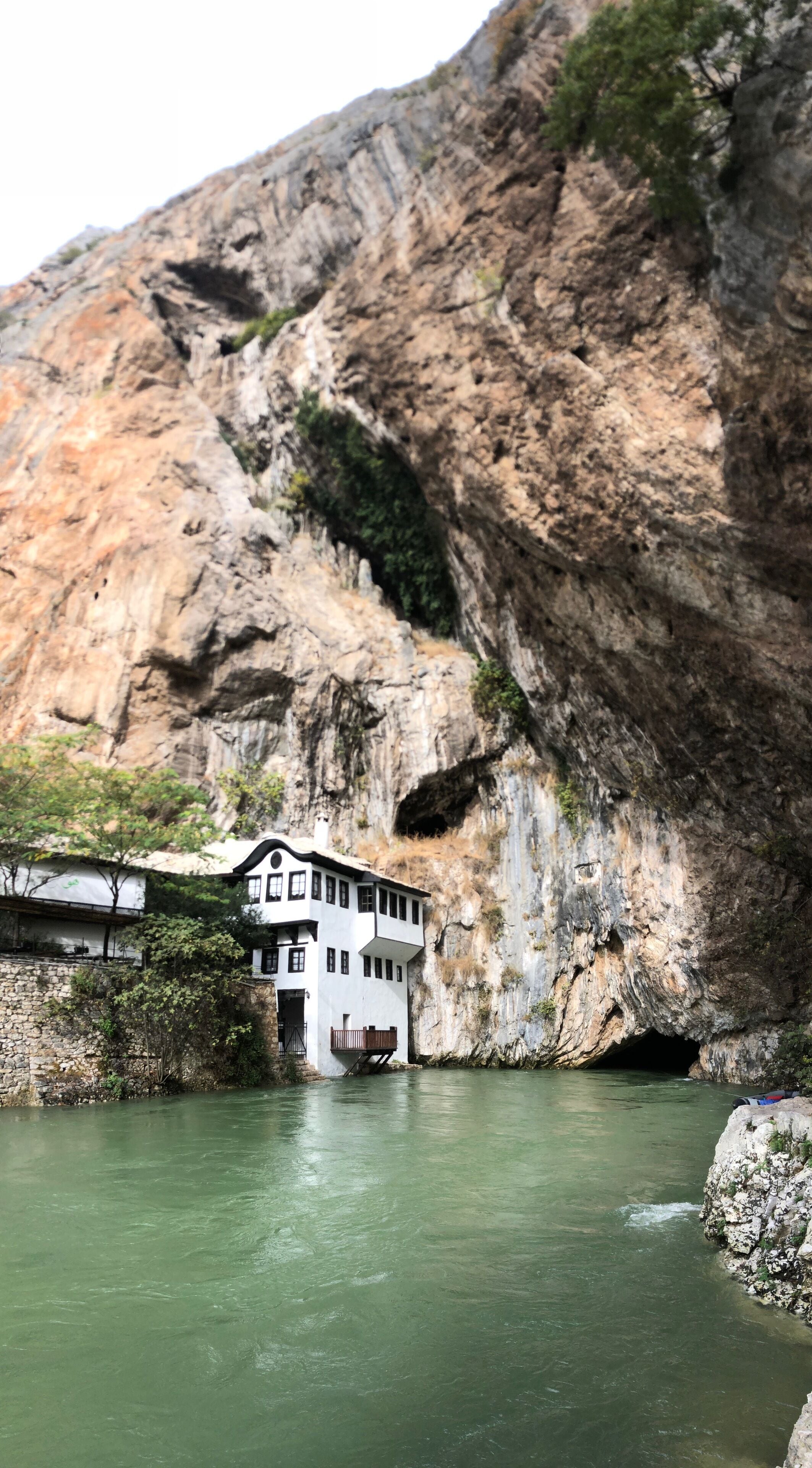 A white building with a balcony overlooking a river, set against a rocky cliff.