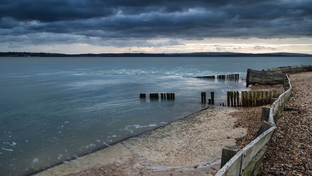 Une plage avec des poteaux en bois plantés dans l’eau, une plage de galets et un ciel nuageux.
