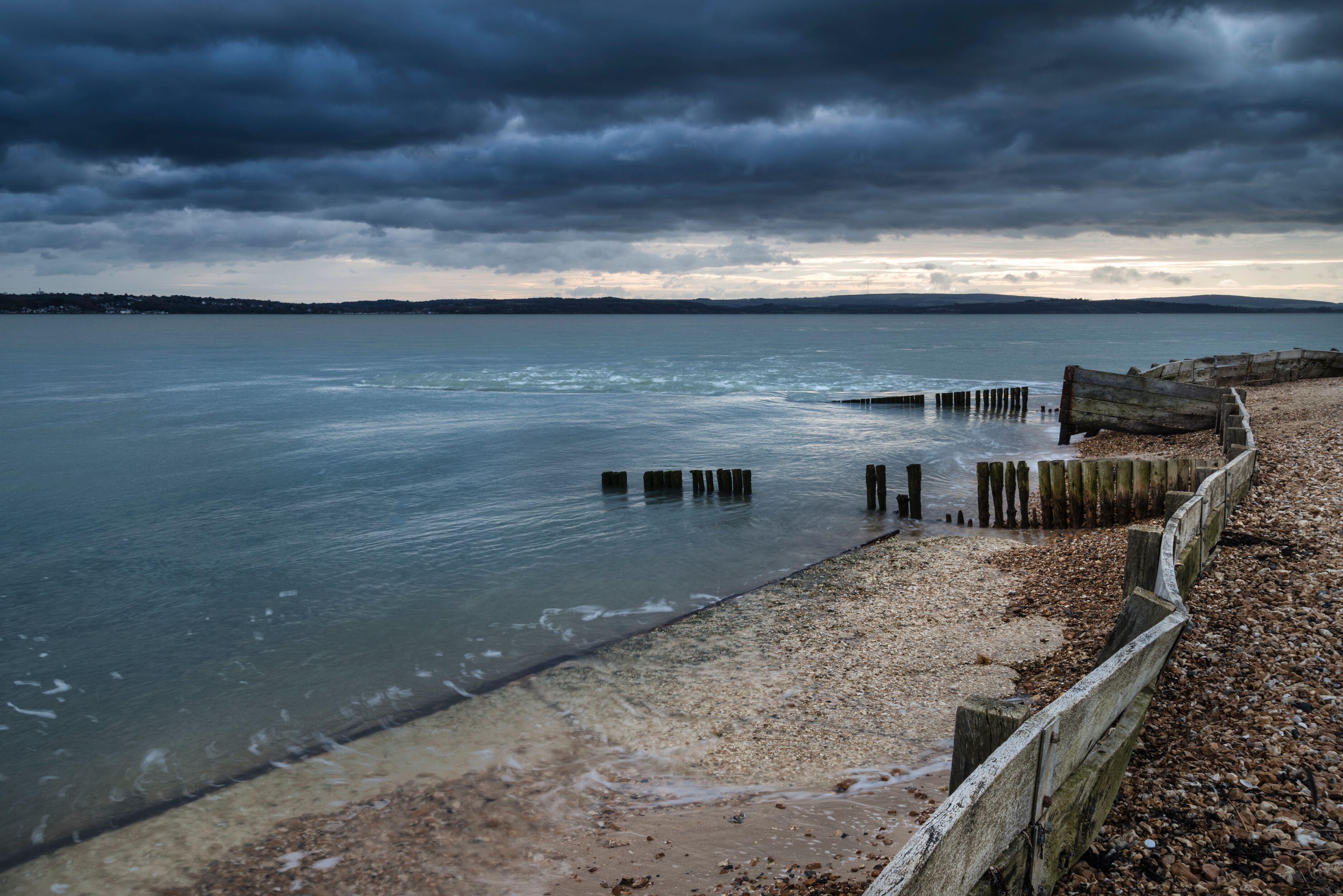 A beach with wooden posts in the water, a pebble beach, and a cloudy sky.