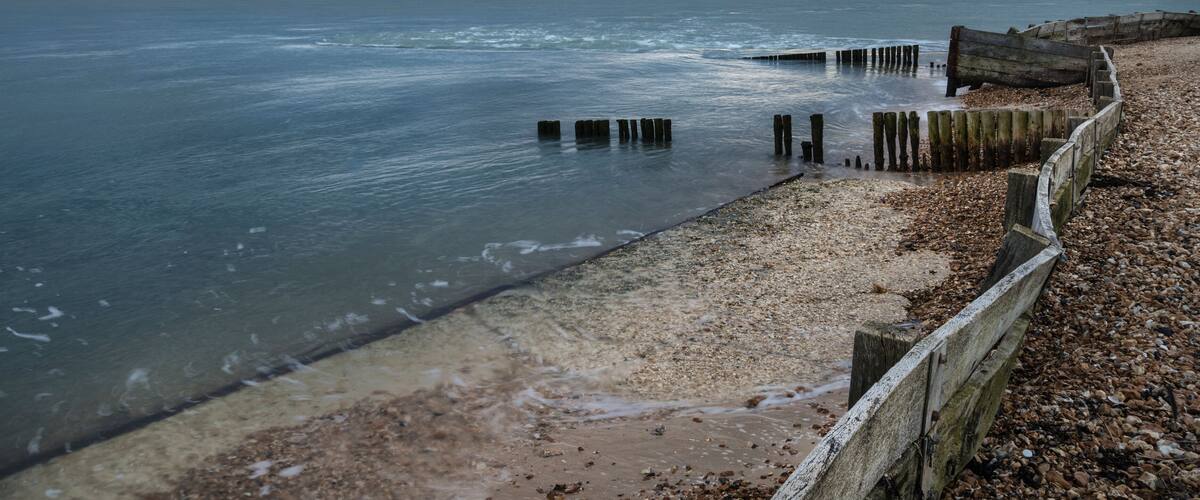 A beach with wooden posts in the water, a pebble beach, and a cloudy sky.