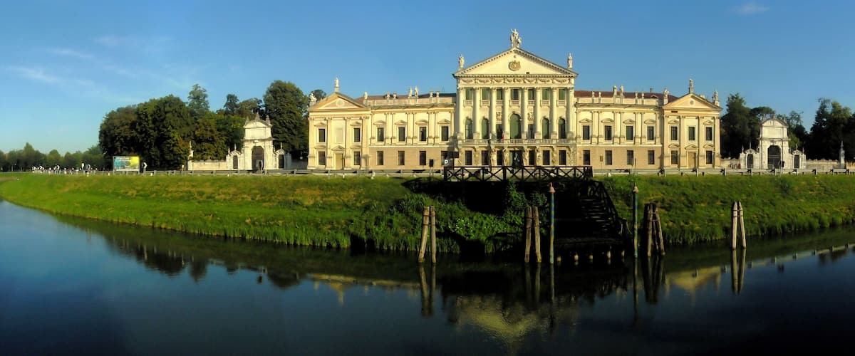 A large classical building with a central dome and symmetrical wings, reflecting in a calm body of water.