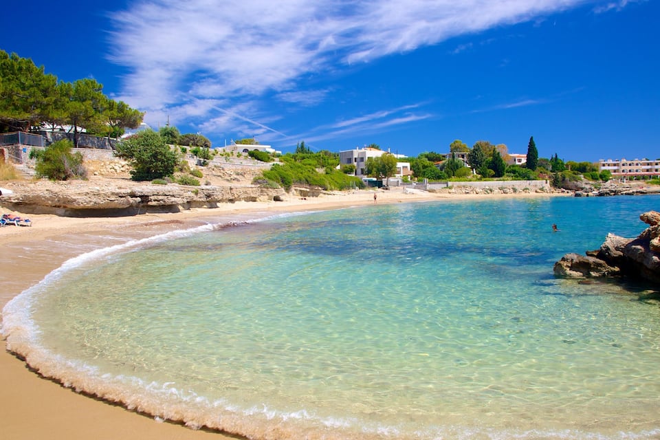 A clear beach with turquoise water, a sandy shore, and buildings in the background.