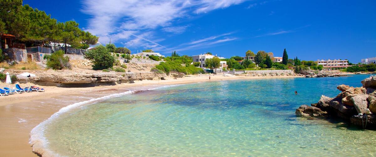 A clear beach with turquoise water, a sandy shore, and buildings in the background.