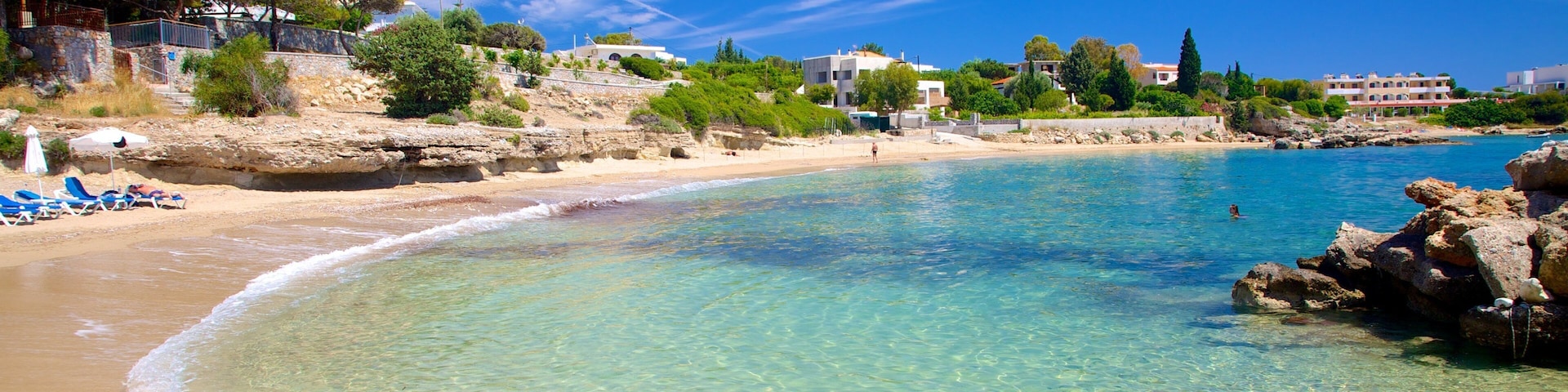 A clear beach with turquoise water, a sandy shore, and buildings in the background.