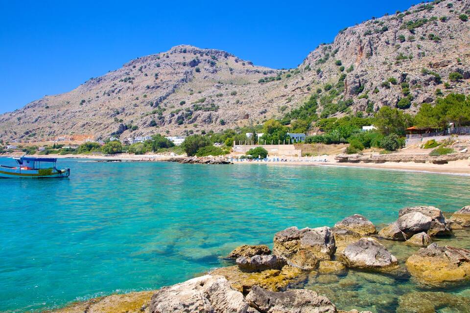 A clear turquoise sea with a boat, rocky shoreline, and a mountainous backdrop.