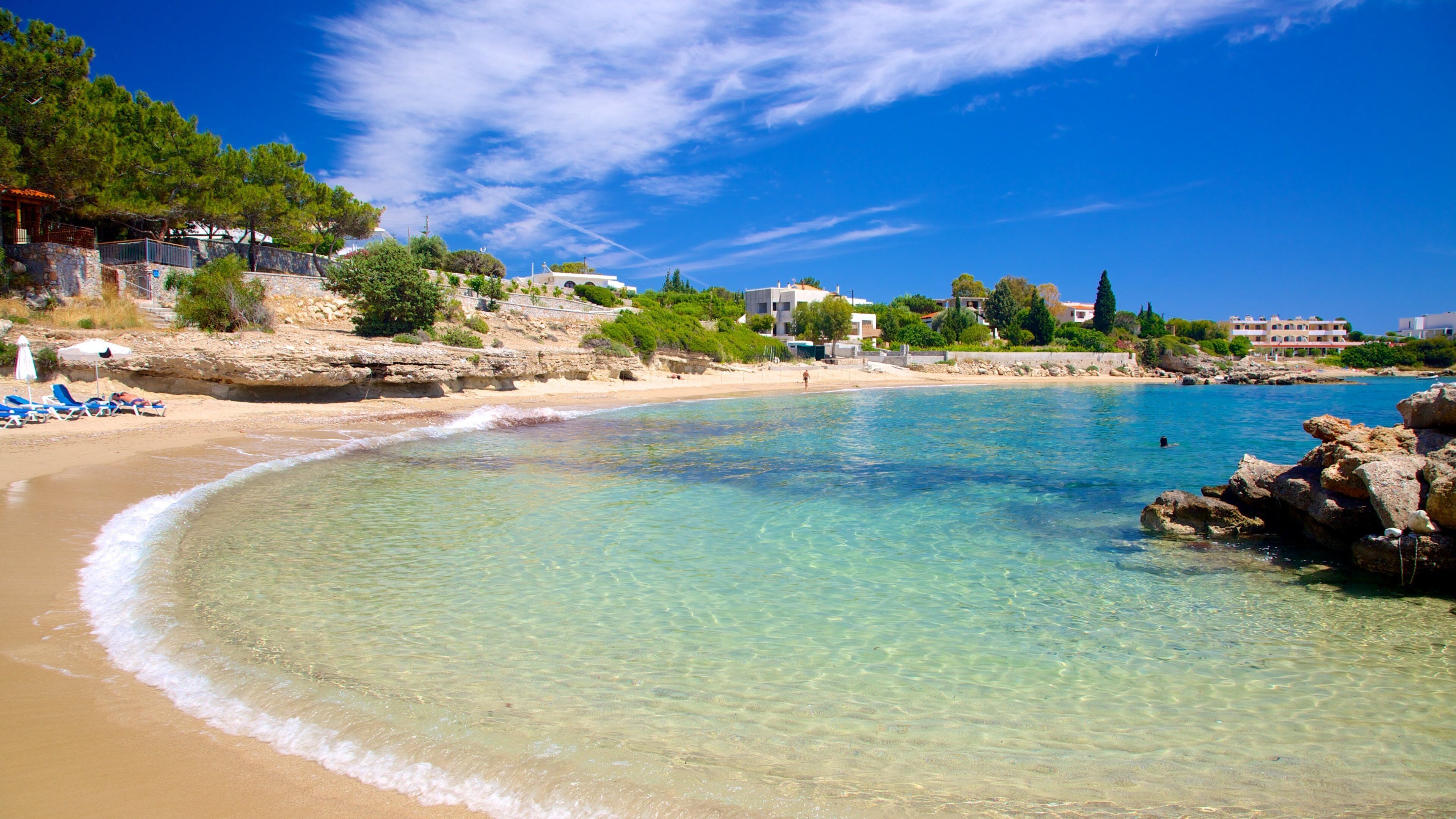 A clear turquoise beach with a rocky shoreline, surrounded by buildings and greenery.