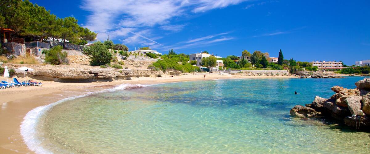A clear turquoise beach with a rocky shoreline, surrounded by buildings and greenery.