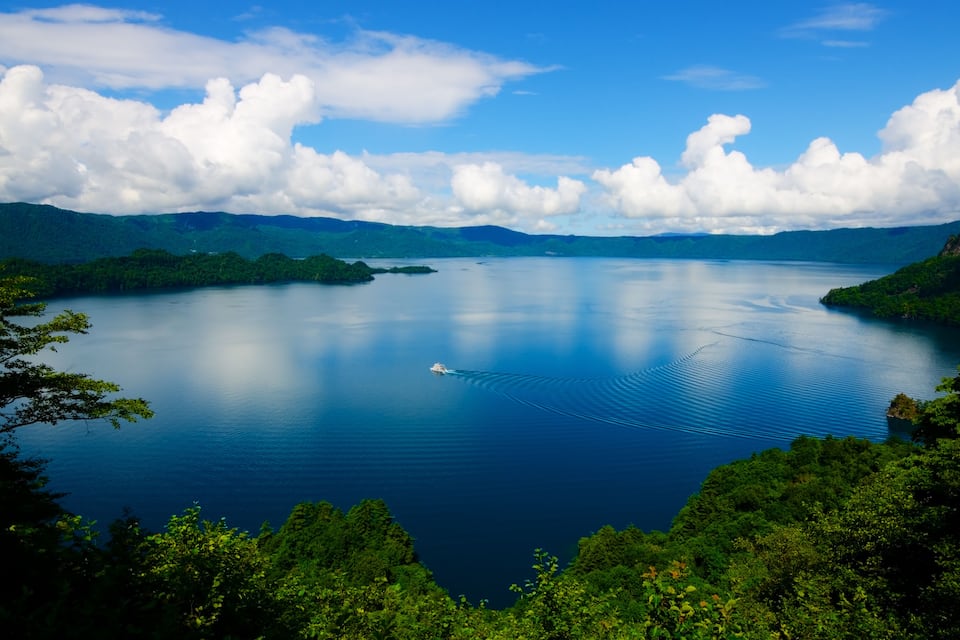 A large calm lake surrounded by lush green hills and a clear blue sky with fluffy clouds.