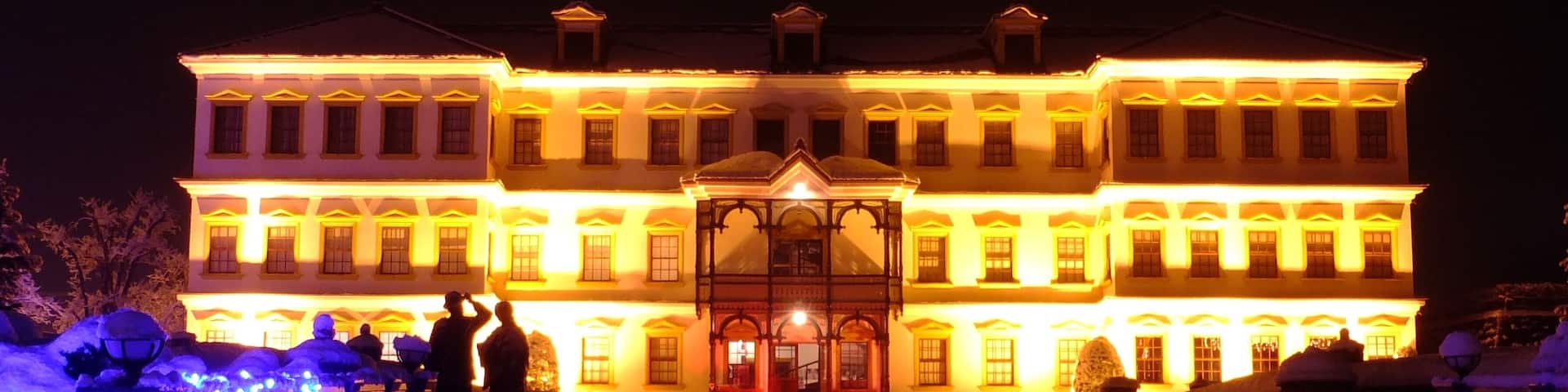 A large, illuminated building with a central entrance and multiple windows, surrounded by snow and decorative lights.