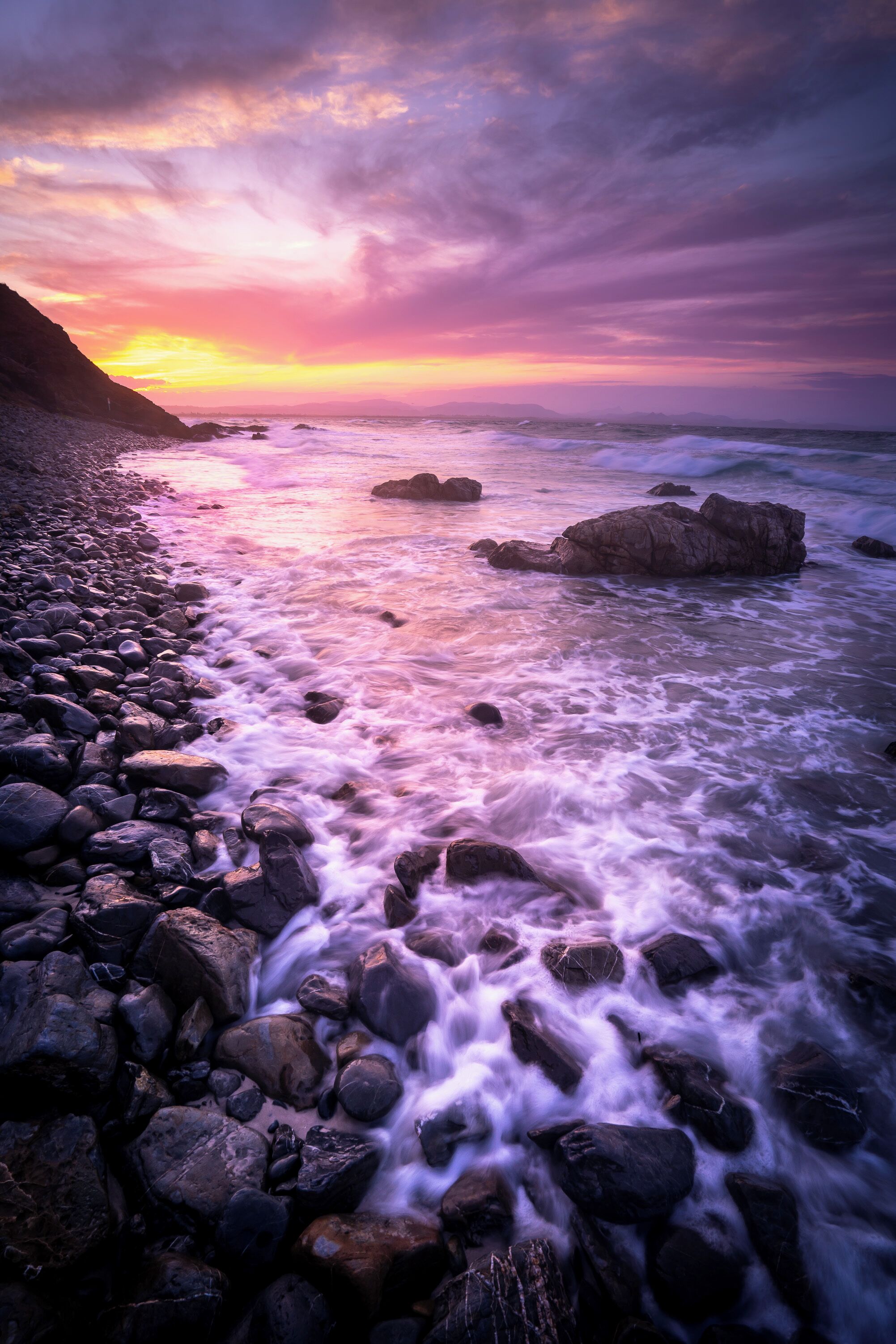 A rocky shoreline with waves crashing against the rocks under a colorful sunset sky.