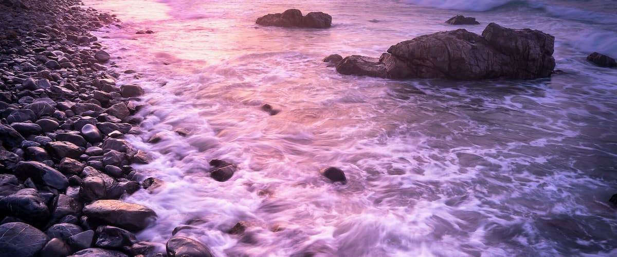 A rocky shoreline with waves crashing against the rocks under a colorful sunset sky.