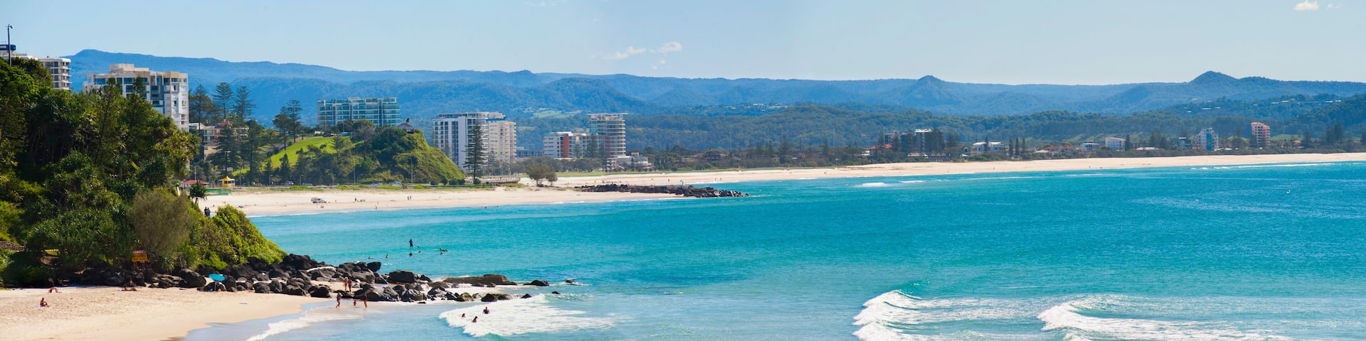 A coastal beach with clear turquoise waters, sandy shore, and a backdrop of mountains and buildings.