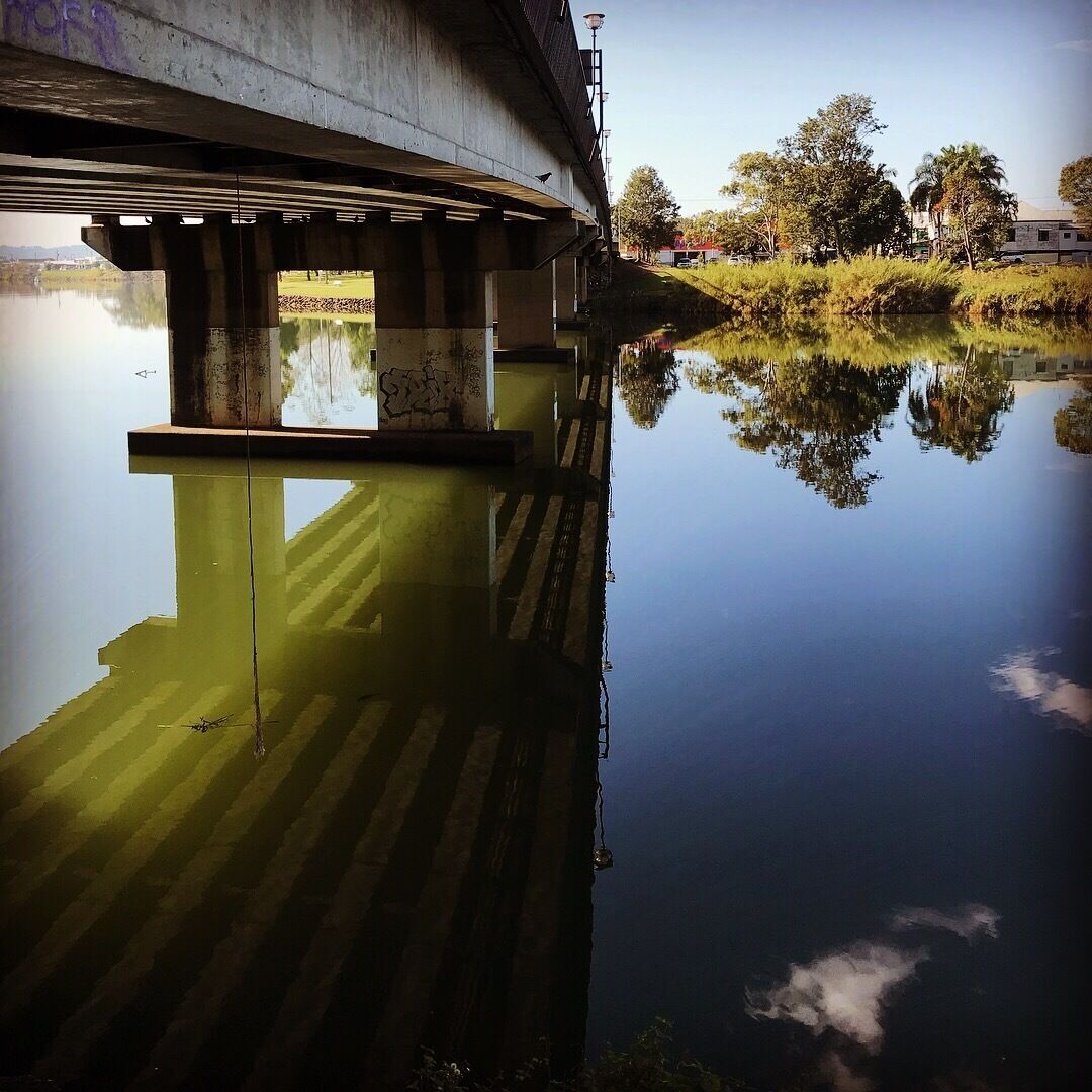 Uma ponte sobre um rio calmo, com reflexos nítidos de árvores e um edifício.