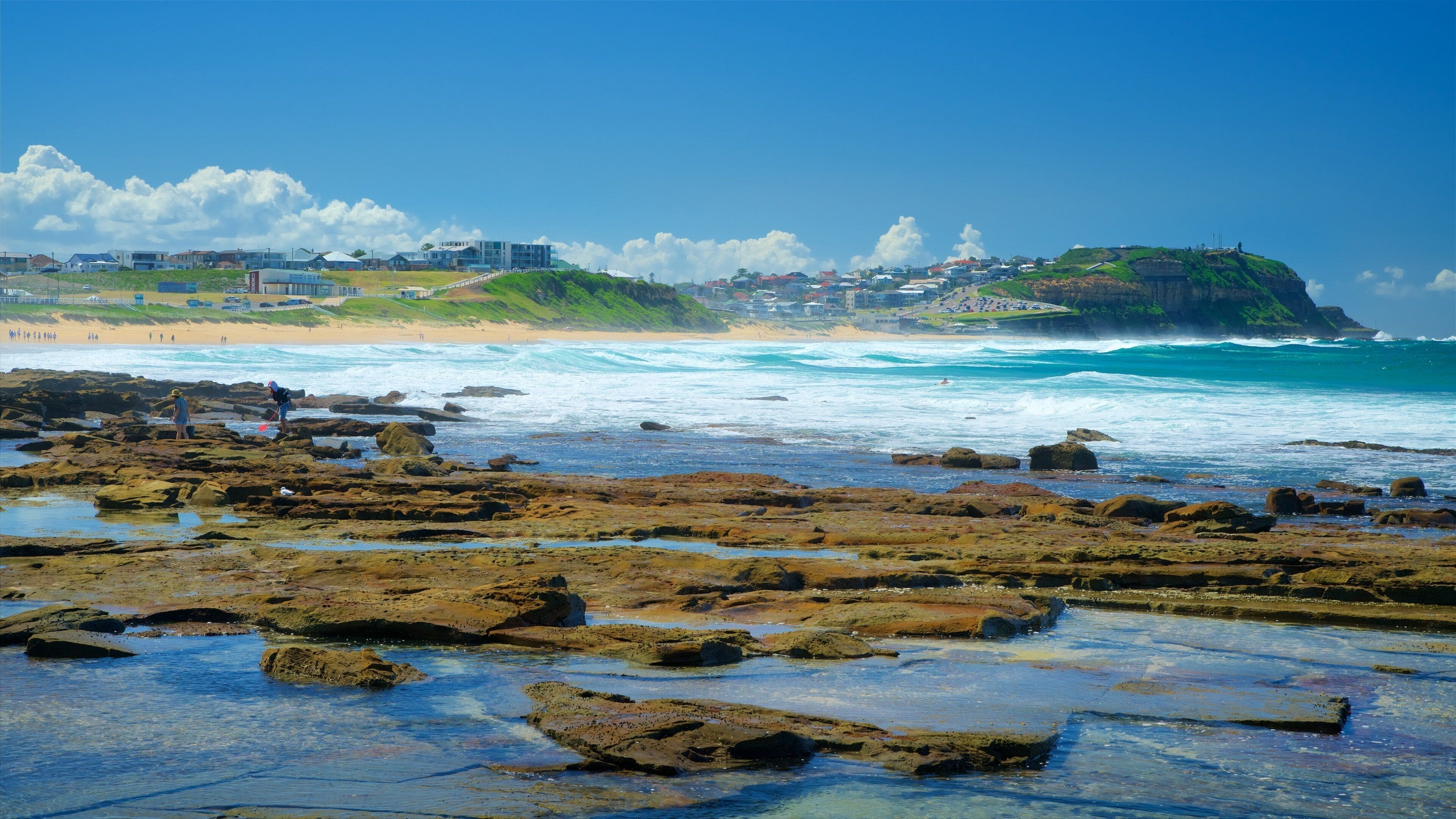 A rocky shoreline with waves crashing onto the beach, a distant town with buildings, and a green hill with a lighthouse.