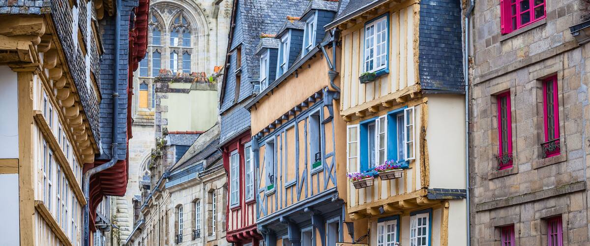 A narrow street lined with colorful half-timbered buildings, a Gothic church, and a stone building with pink shutters.