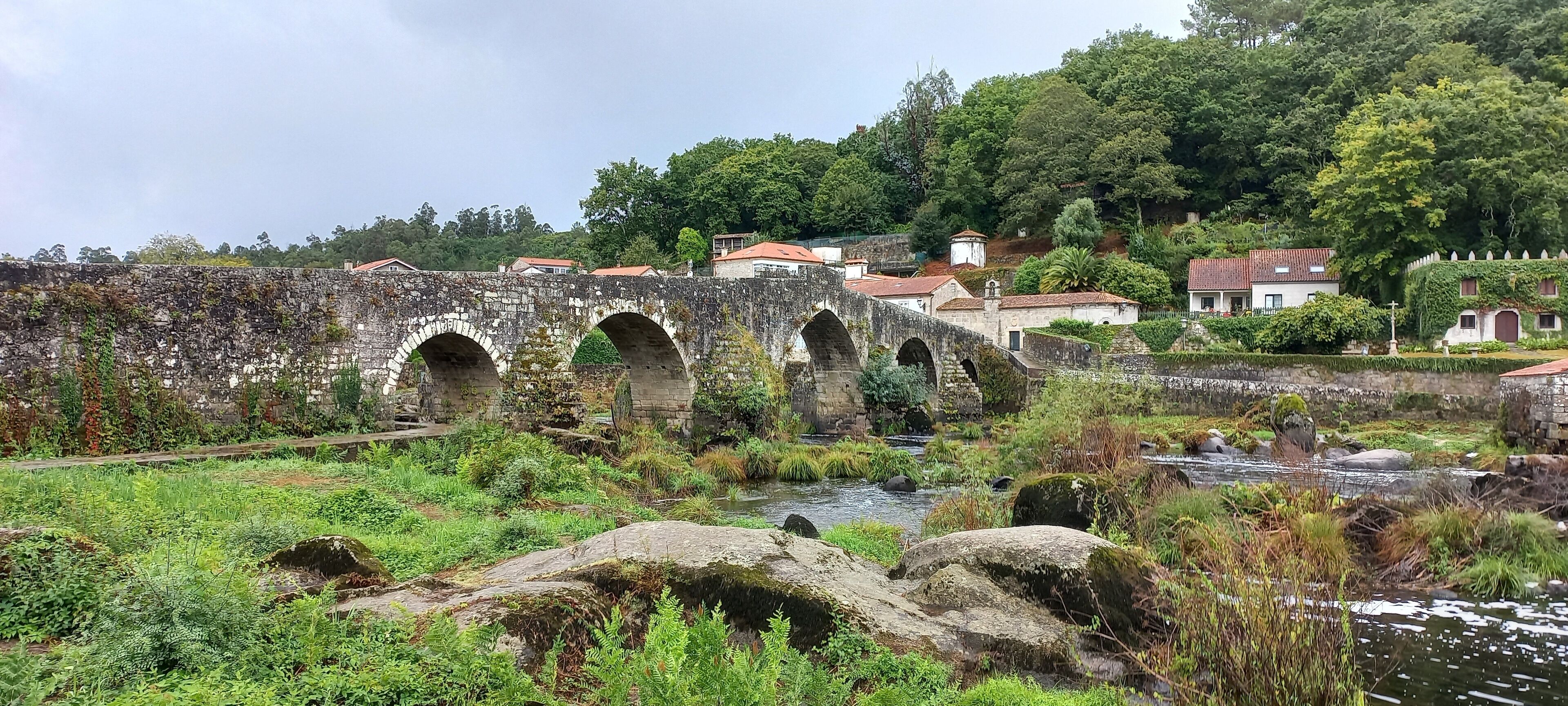 A stone bridge with three arches over a river, surrounded by lush greenery and houses with red roofs.