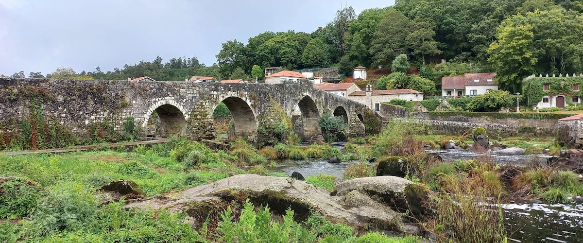 A stone bridge with three arches over a river, surrounded by lush greenery and houses with red roofs.