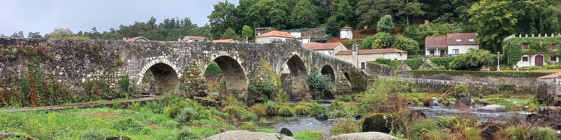 A stone bridge with three arches over a river, surrounded by lush greenery and houses with red roofs.