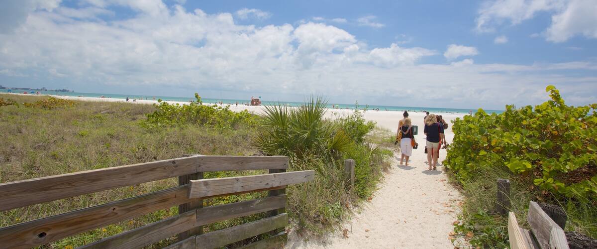 A sandy beach path with people walking, a wooden fence, and a clear blue sky with clouds.