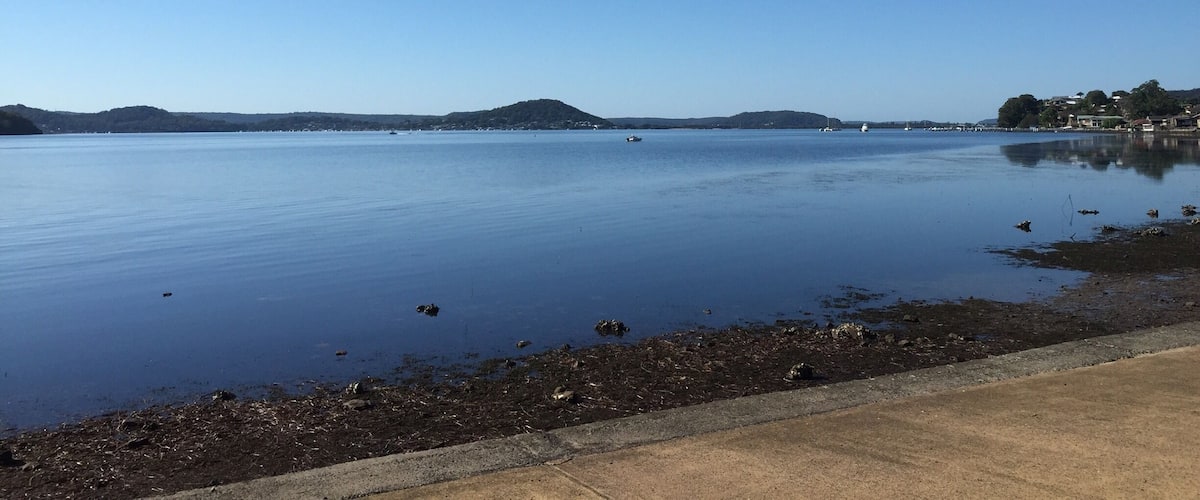 A calm lake with a clear blue sky, a distant hill, and a shoreline with buildings.