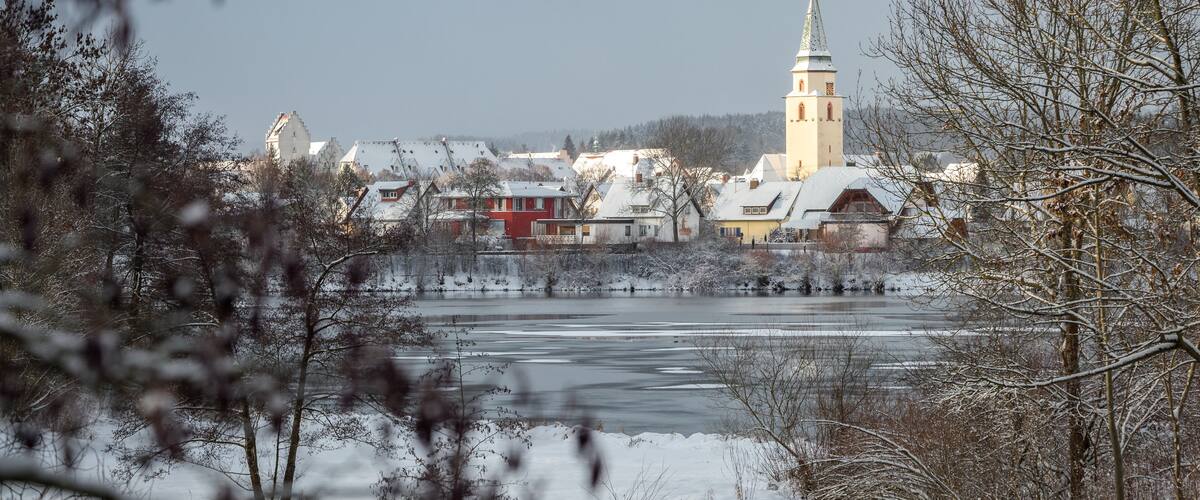 A winter scene with a frozen river, snow-covered trees, and a church with a tall spire.