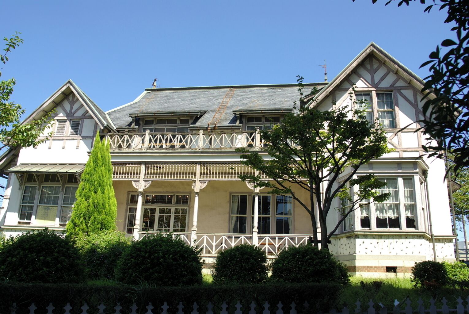A large, historic house with a prominent balcony, a tall coniferous tree, and a well-maintained garden.