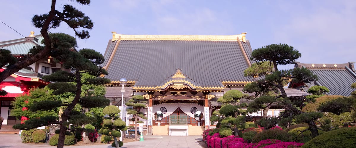 A traditional Japanese building with a tiled roof, surrounded by well-manicured trees and vibrant pink flowers.