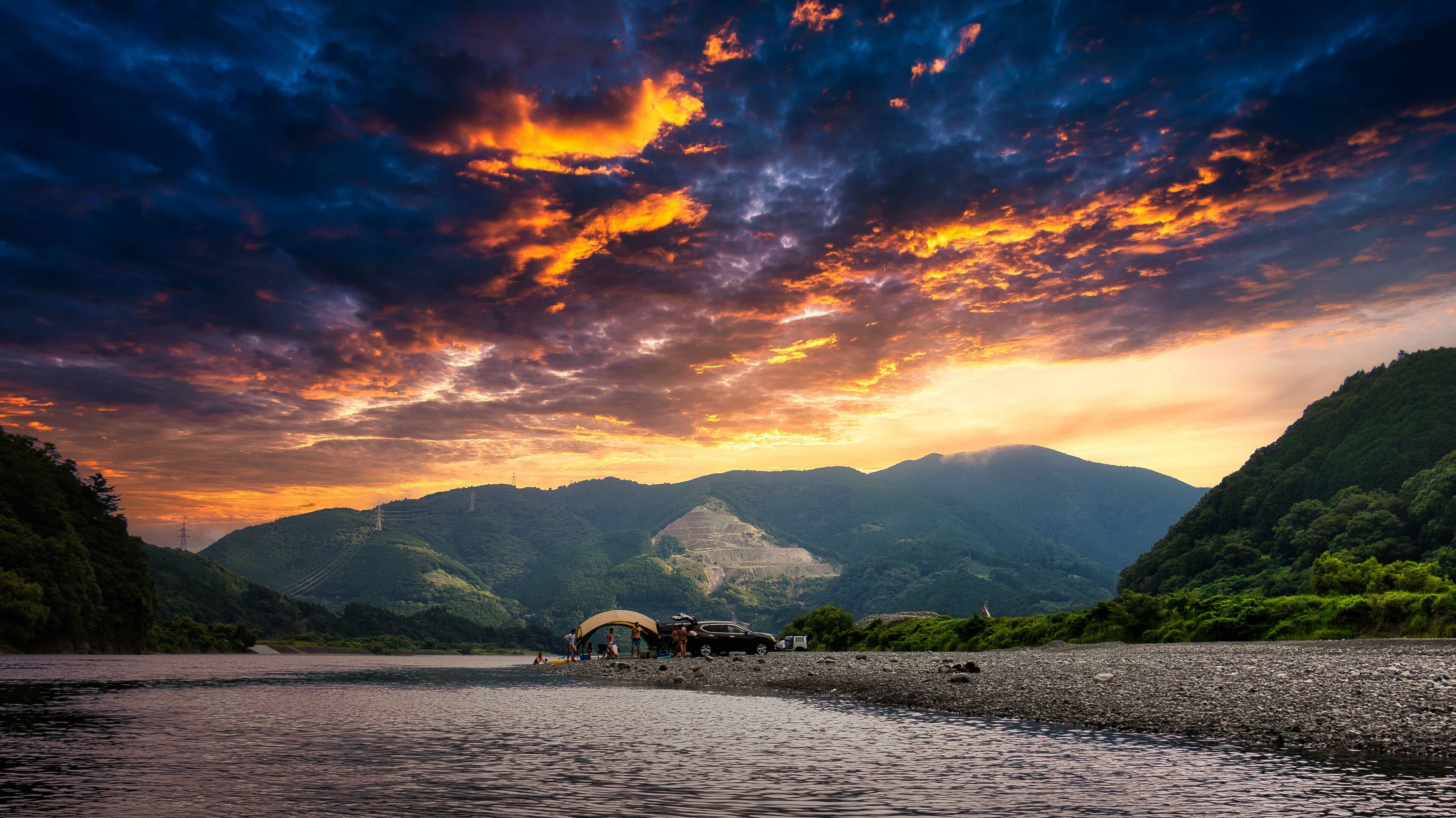 A river with a rocky shore, a group of people, and a car parked near a tent.