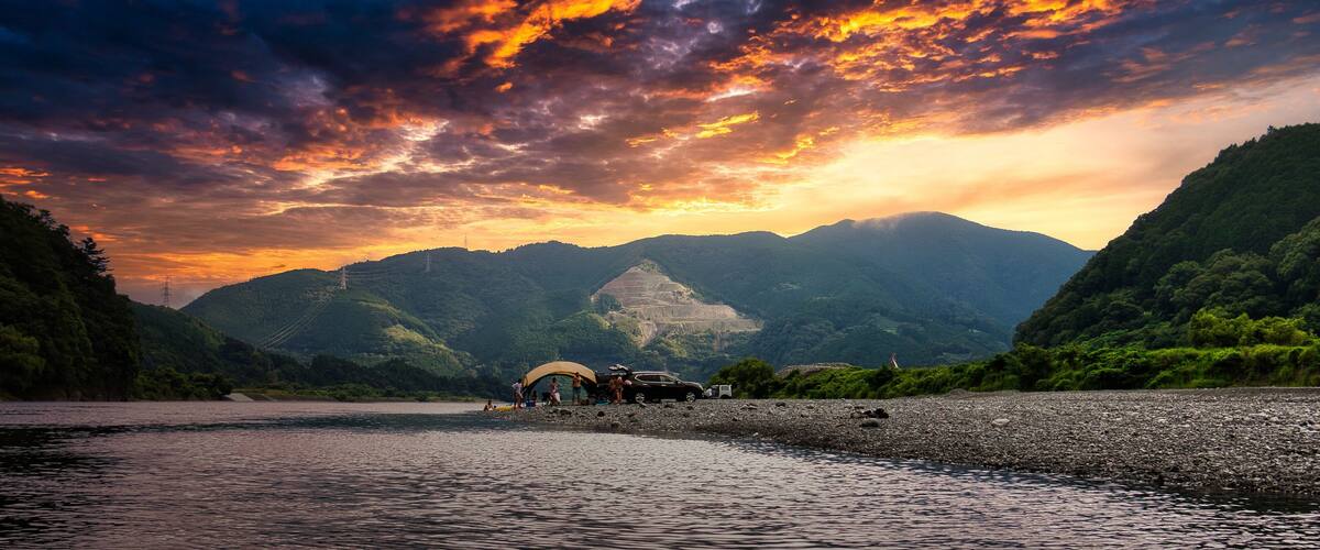 A river with a rocky shore, a group of people, and a car parked near a tent.