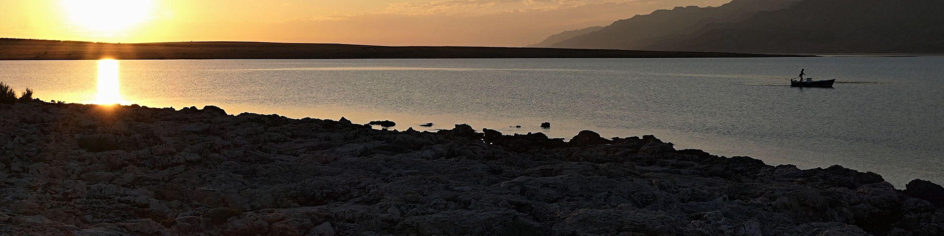 Evening landscape with setting sun, rocky beach silhouette in forefront, bay with single fisherman boat and Velebit mountain in background. Clear skies with some clouds. Location near Razanac, Croatia