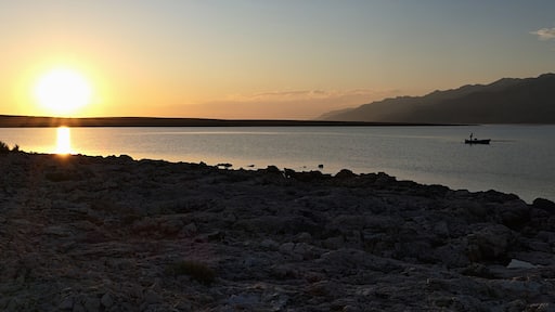 Evening landscape with setting sun, rocky beach silhouette in forefront, bay with single fisherman boat and Velebit mountain in background. Clear skies with some clouds. Location near Razanac, Croatia
