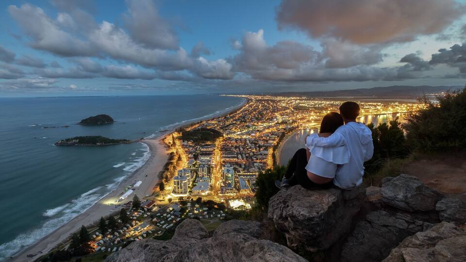 A coastal cityscape at dusk with a view of the beach, a rocky outcrop, and a distant island.