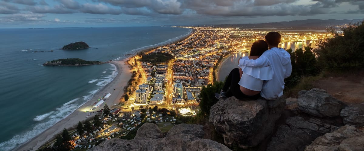 A coastal cityscape at dusk with a view of the beach, a rocky outcrop, and a distant island.