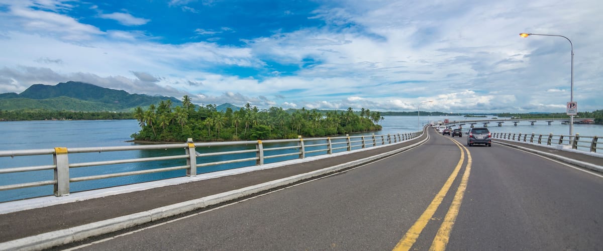 A bridge with a guardrail, a road, and a body of water with mountains in the background.
