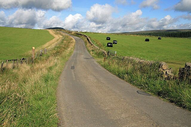 A rural road with a stone wall on one side and green fields on the other.