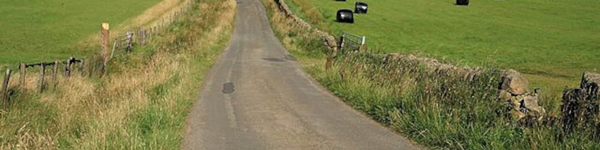 A rural road with a stone wall on one side and green fields on the other.