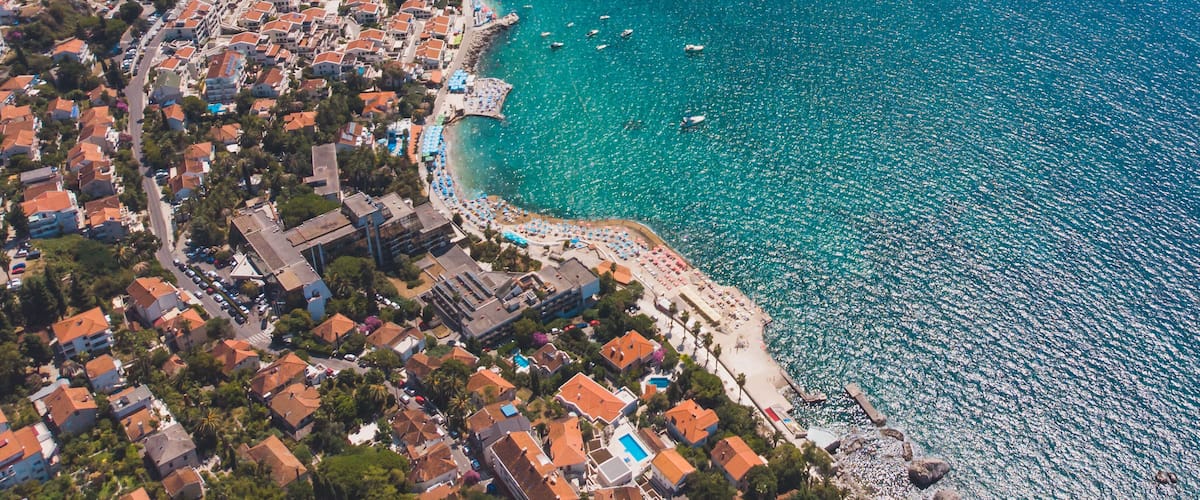 An aerial view of a coastal town with a clear blue sea, a sandy beach, and densely packed buildings with red roofs.