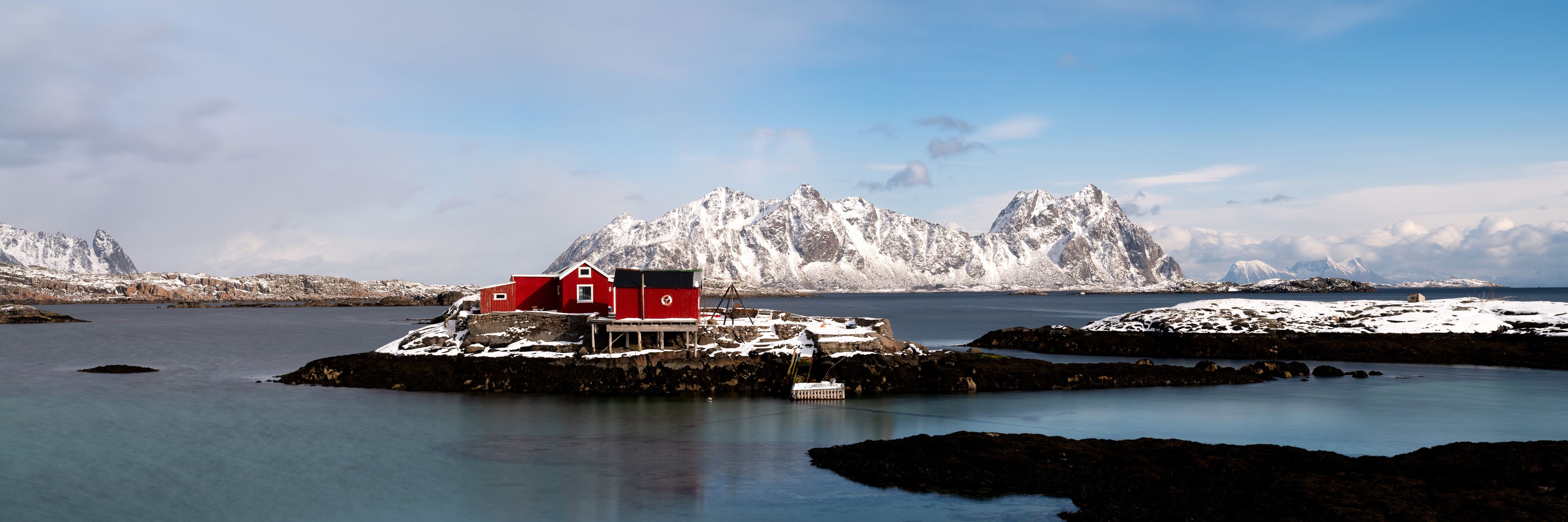 A red wooden house on a small island with a fjord and snow-covered mountains in the background.