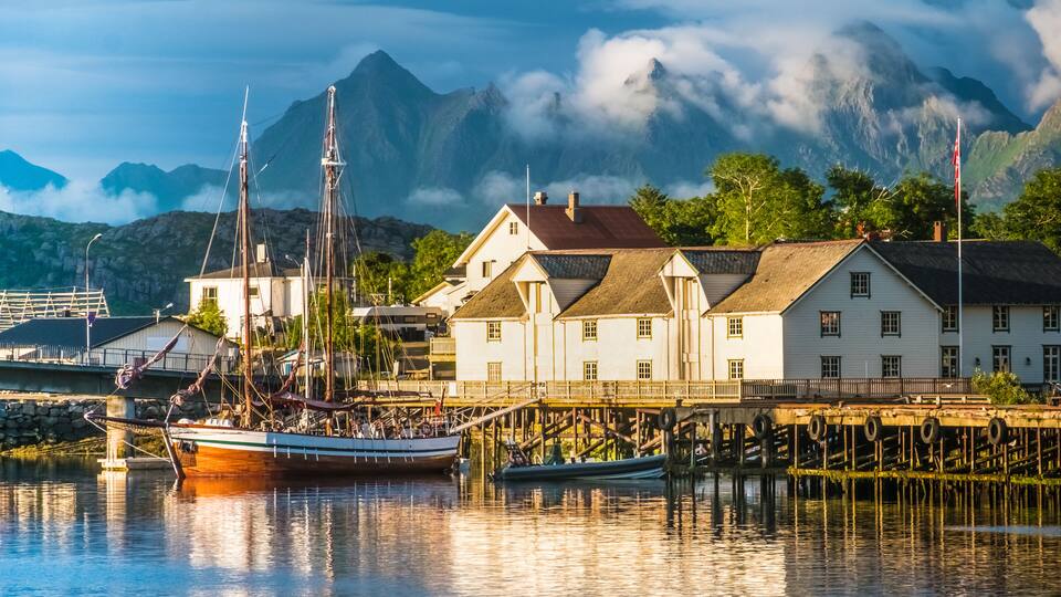 A harbor with a sailing ship, white buildings, and mountains in the background.