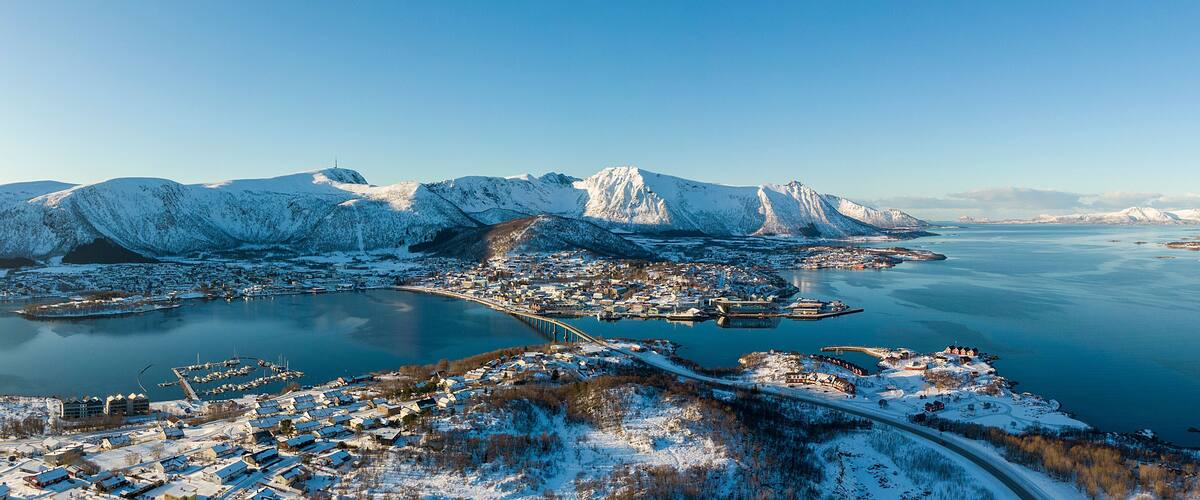 A snowy coastal town with mountains in the background and a bridge connecting the town to the mainland.