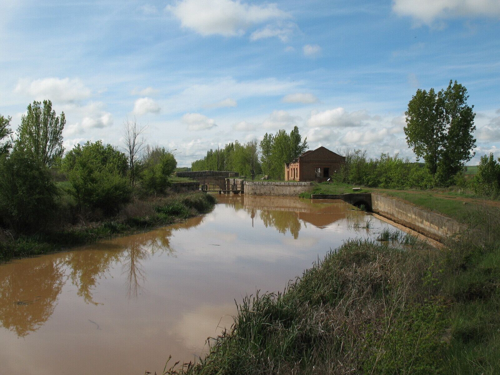 A calm river with a stone bridge, surrounded by greenery and a small building.