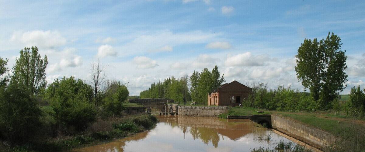 A calm river with a stone bridge, surrounded by greenery and a small building.