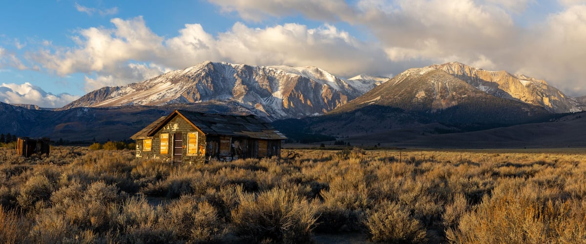 A rustic cabin in a dry grass field with snow-capped mountains in the background.