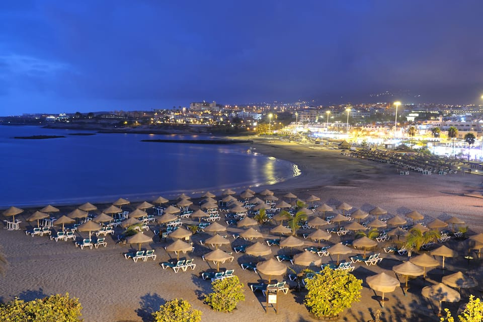 A beach with rows of sun loungers and umbrellas, a calm sea, and a cityscape in the background.