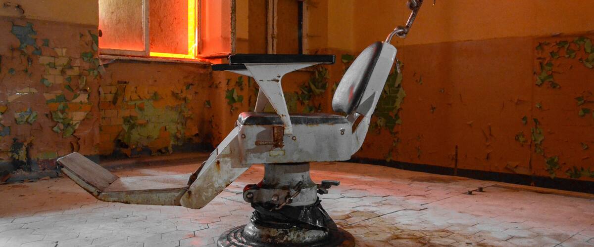 An abandoned barber chair in a dilapidated room with peeling walls and a broken window.