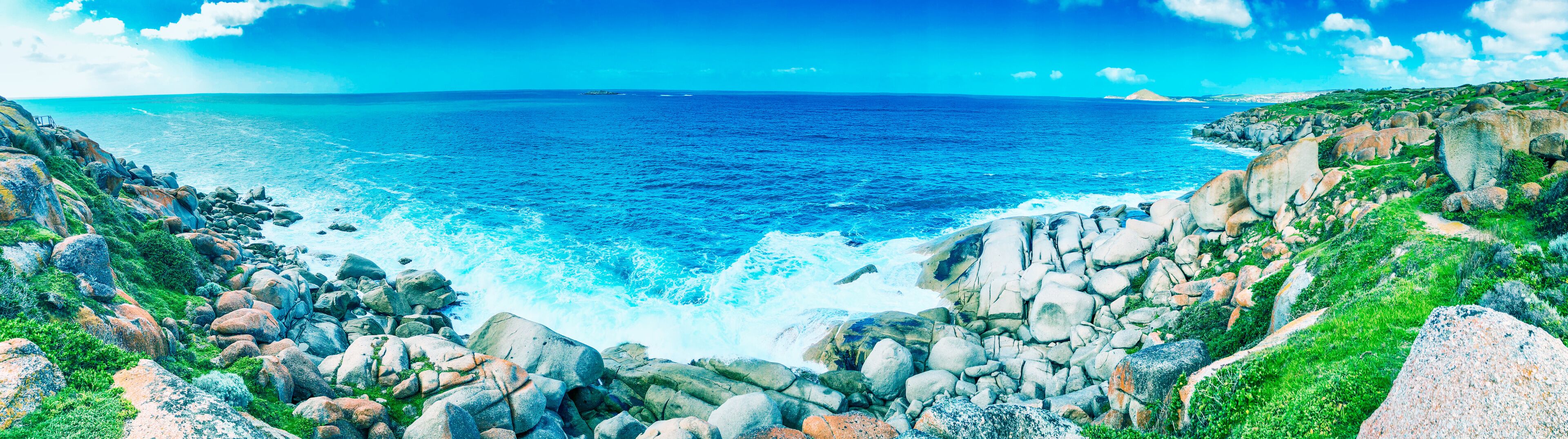 A rocky coastline with waves crashing against the shore, a clear blue sky, and distant landforms.