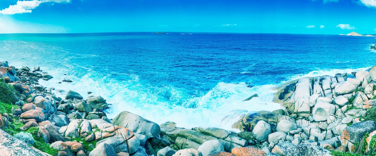 A rocky coastline with waves crashing against the shore, a clear blue sky, and distant landforms.