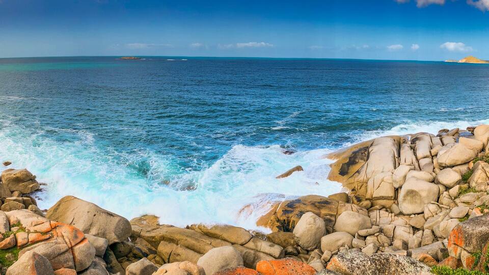 A rocky coastline with waves crashing against the shore, a clear blue sky, and distant landforms.