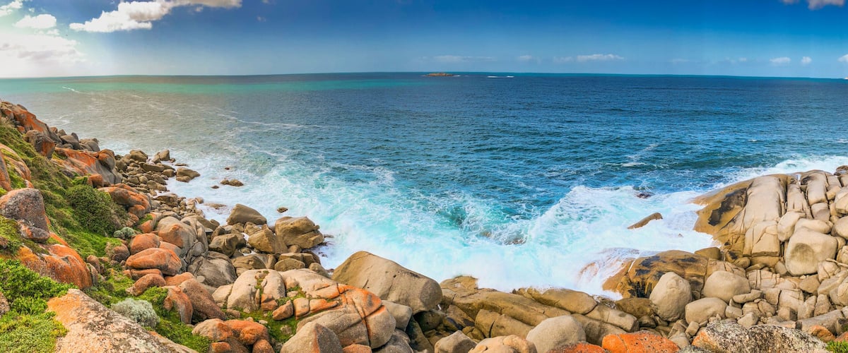 A rocky coastline with waves crashing against the shore, a clear blue sky, and distant landforms.